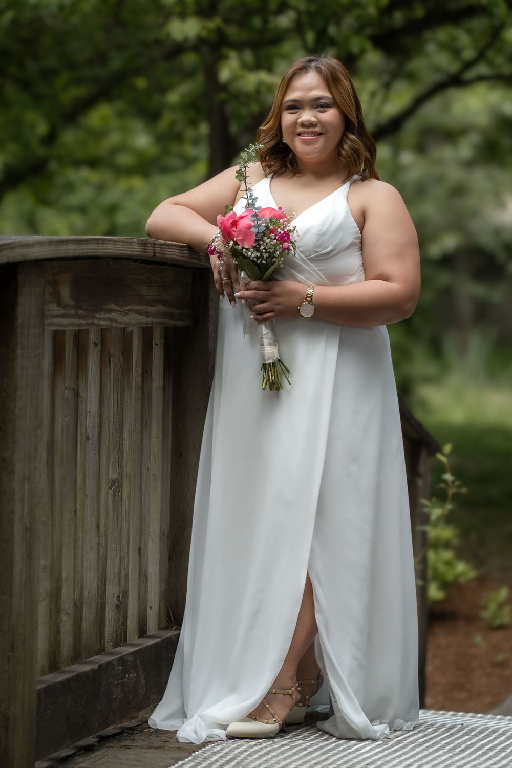 Bride in white gown holding pink bouquet poses on wooden bridge surrounded by lush green trees.