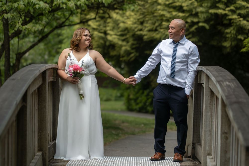 Bride and groom holding hands on a wooden bridge surrounded by lush green trees in an outdoor wedding photo.
