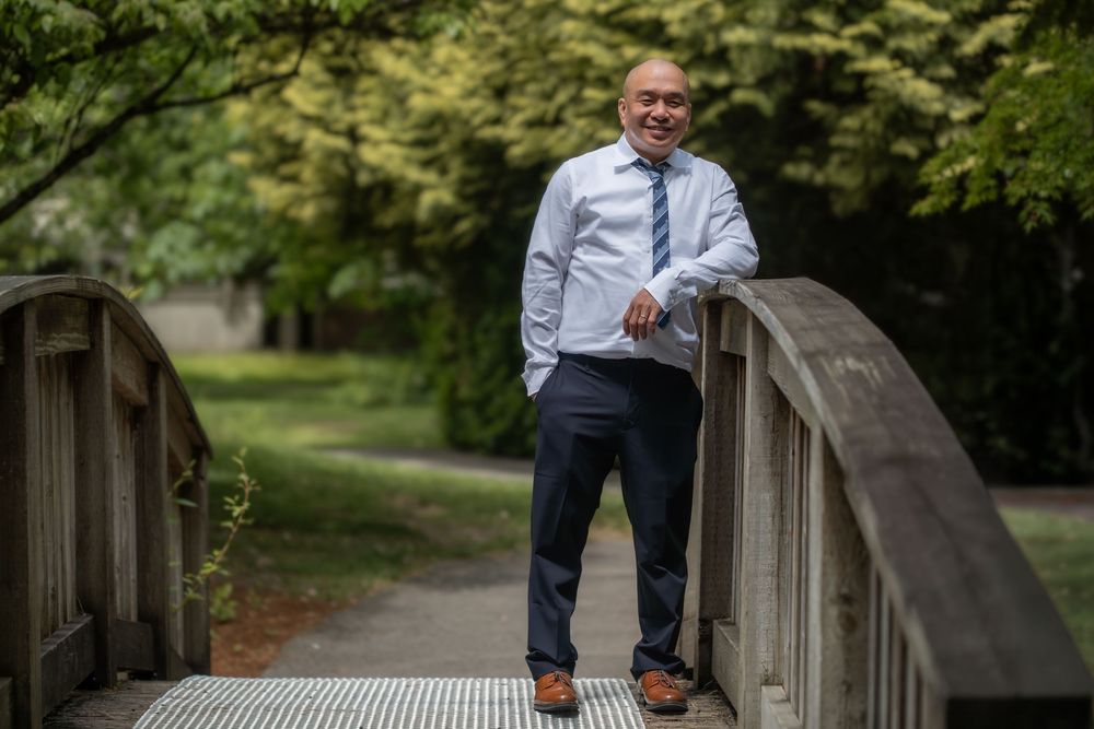 Professional man in dress shirt and slacks smiling on a wooden bridge surrounded by lush green trees.