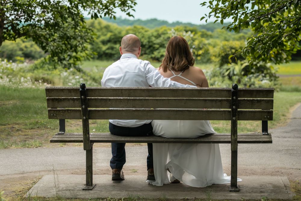 Bride and groom sitting together on a wooden park bench surrounded by lush green trees on their wedding day.