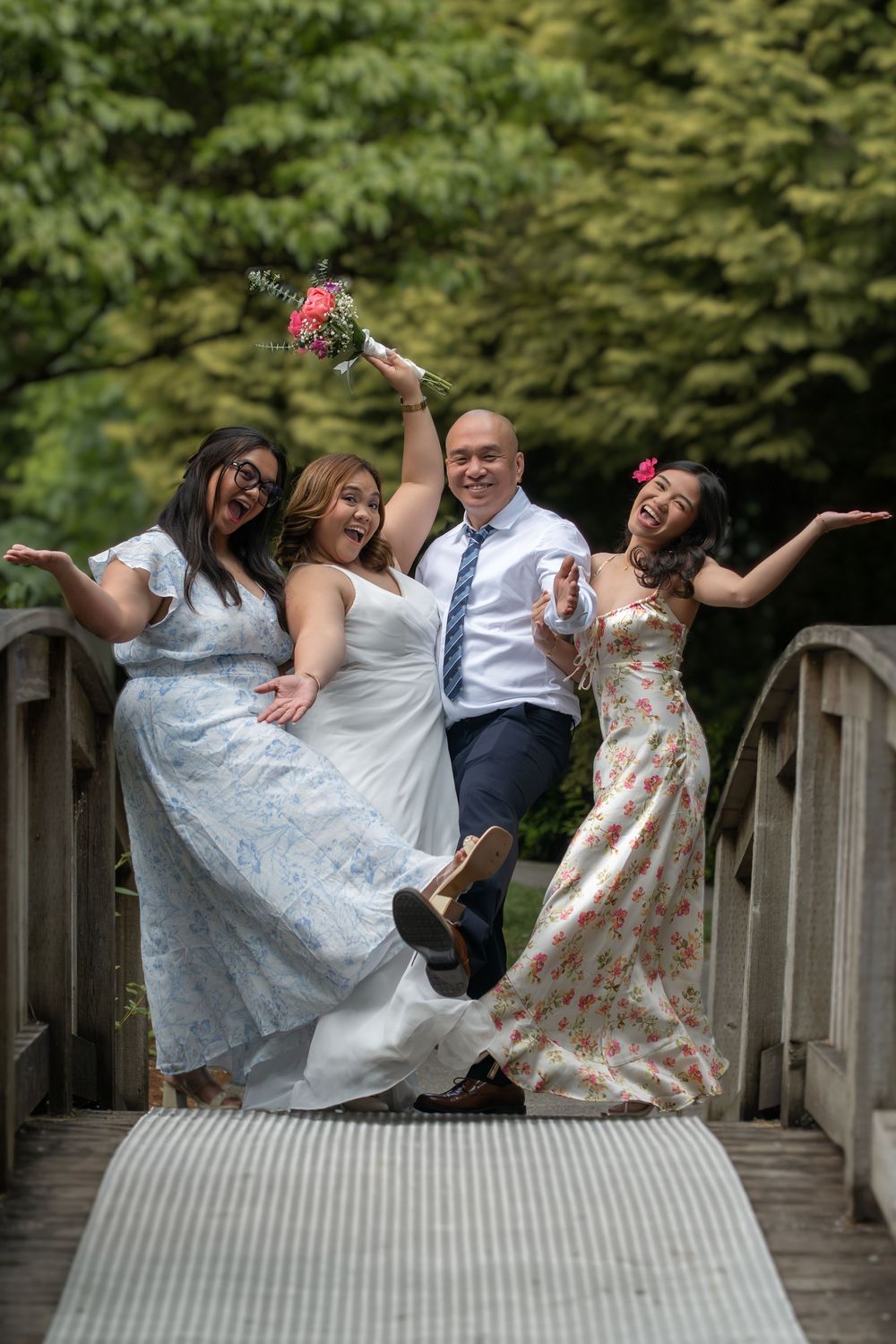 Joyful wedding party of four celebrating on a wooden bridge, bride holding bouquet, surrounded by lush green trees.