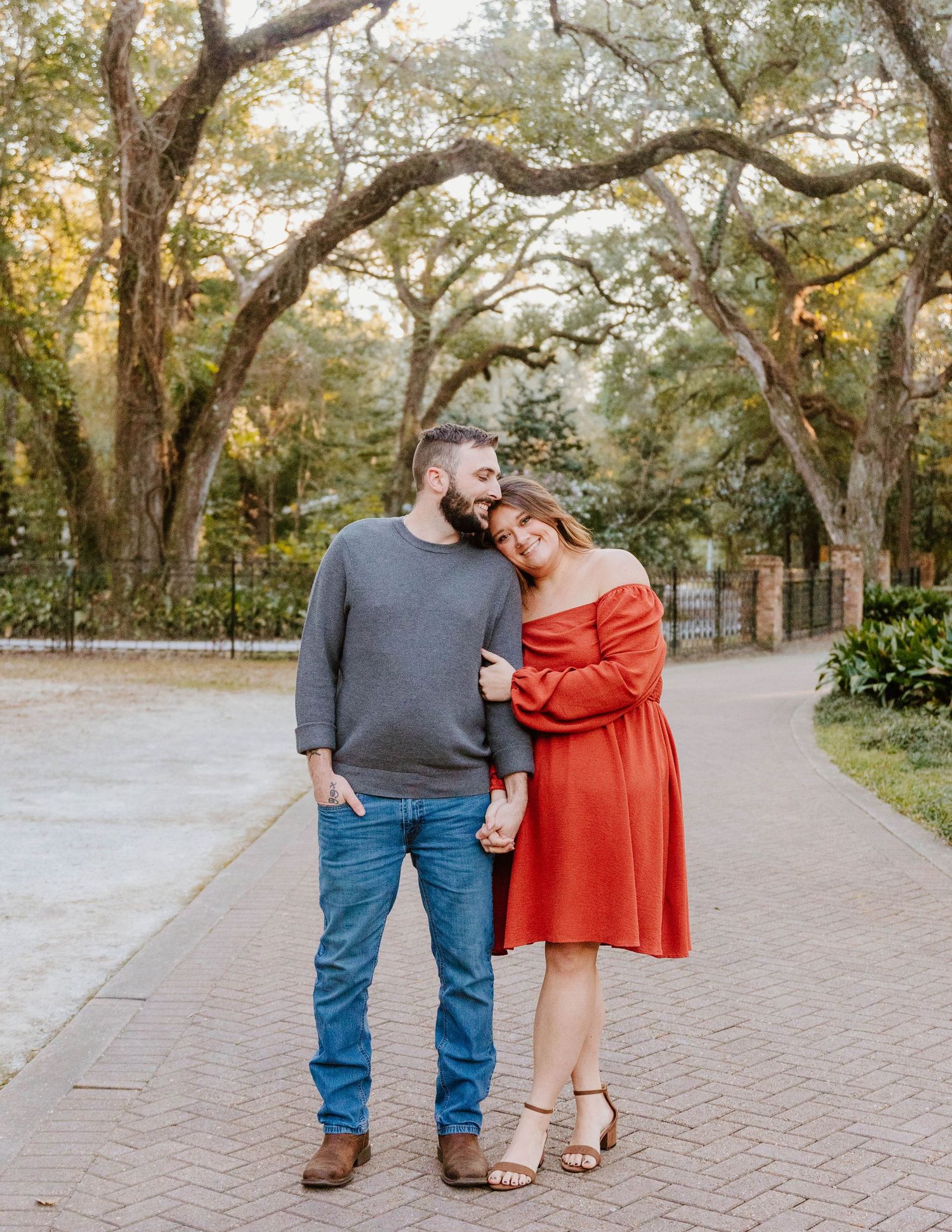 Couple posing on a brick path under oak trees, woman in red off-shoulder dress leans on man in gray sweater and jeans.