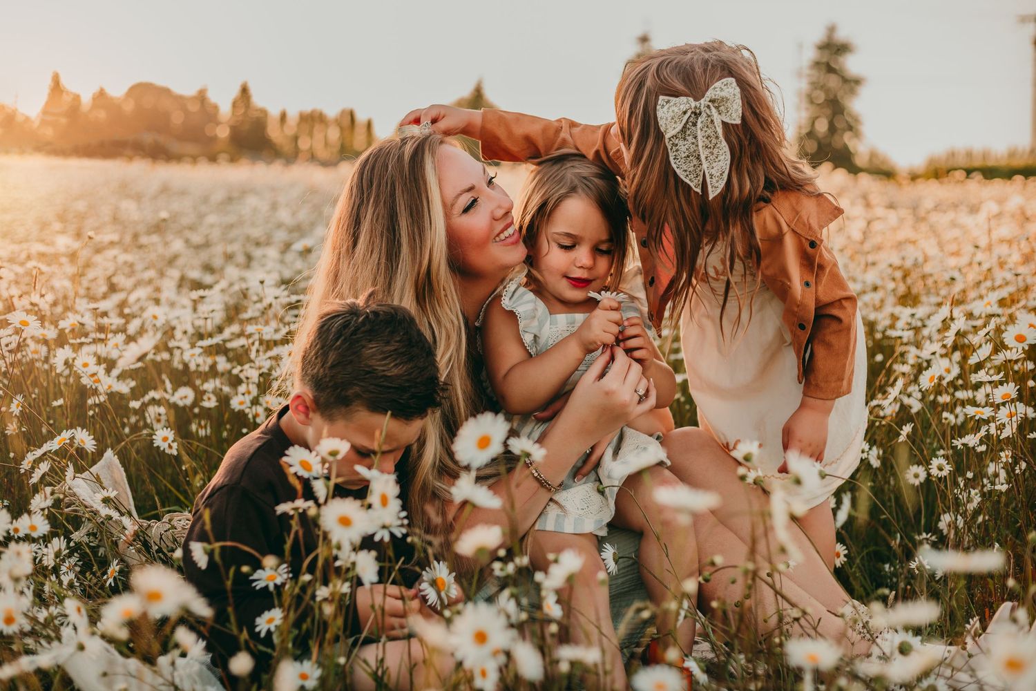 Mother kissing young child in sunny daisy field, surrounded by three children in warm golden light.