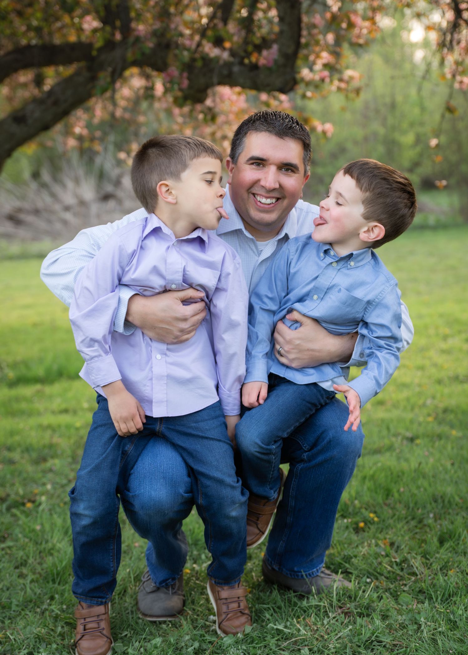 Family of four posing outdoors under blooming spring trees on green grass, parents with two young boys smiling.
