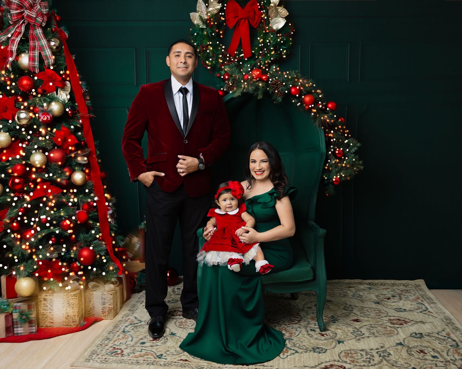Family Christmas portrait with man in red velvet blazer, woman in green gown, and baby in red dress by decorated tree.