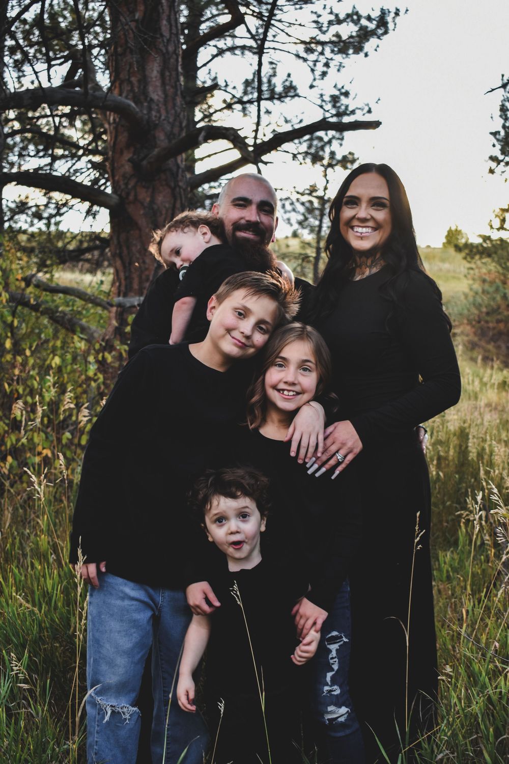 Family of six posing outdoors in matching black outfits among tall grass and pine trees.