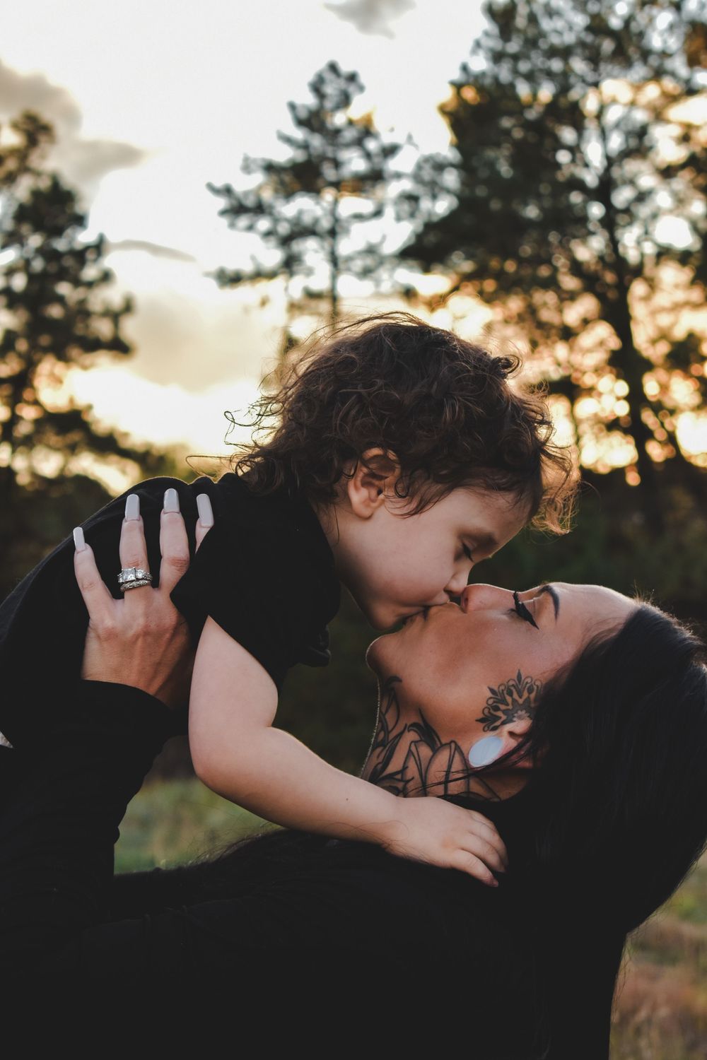 A mother kisses her curly-haired toddler at sunset in a forest setting.