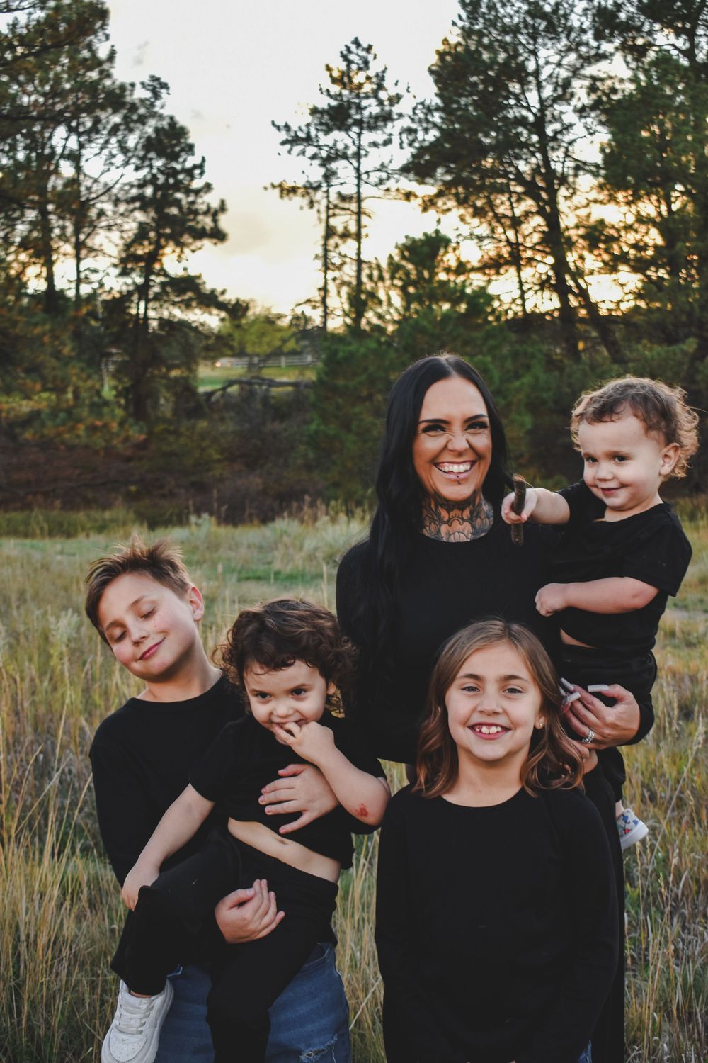 Mother in black dress holding a toddler while posing with three children in a pine forest.