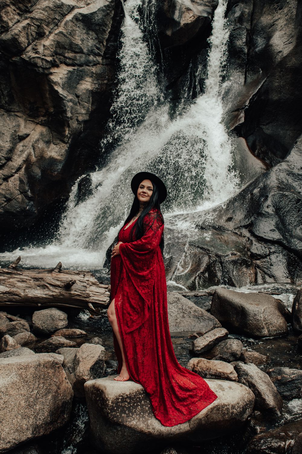 Woman in red dress and black hat poses on rocks before a cascading waterfall in a dramatic outdoor portrait.