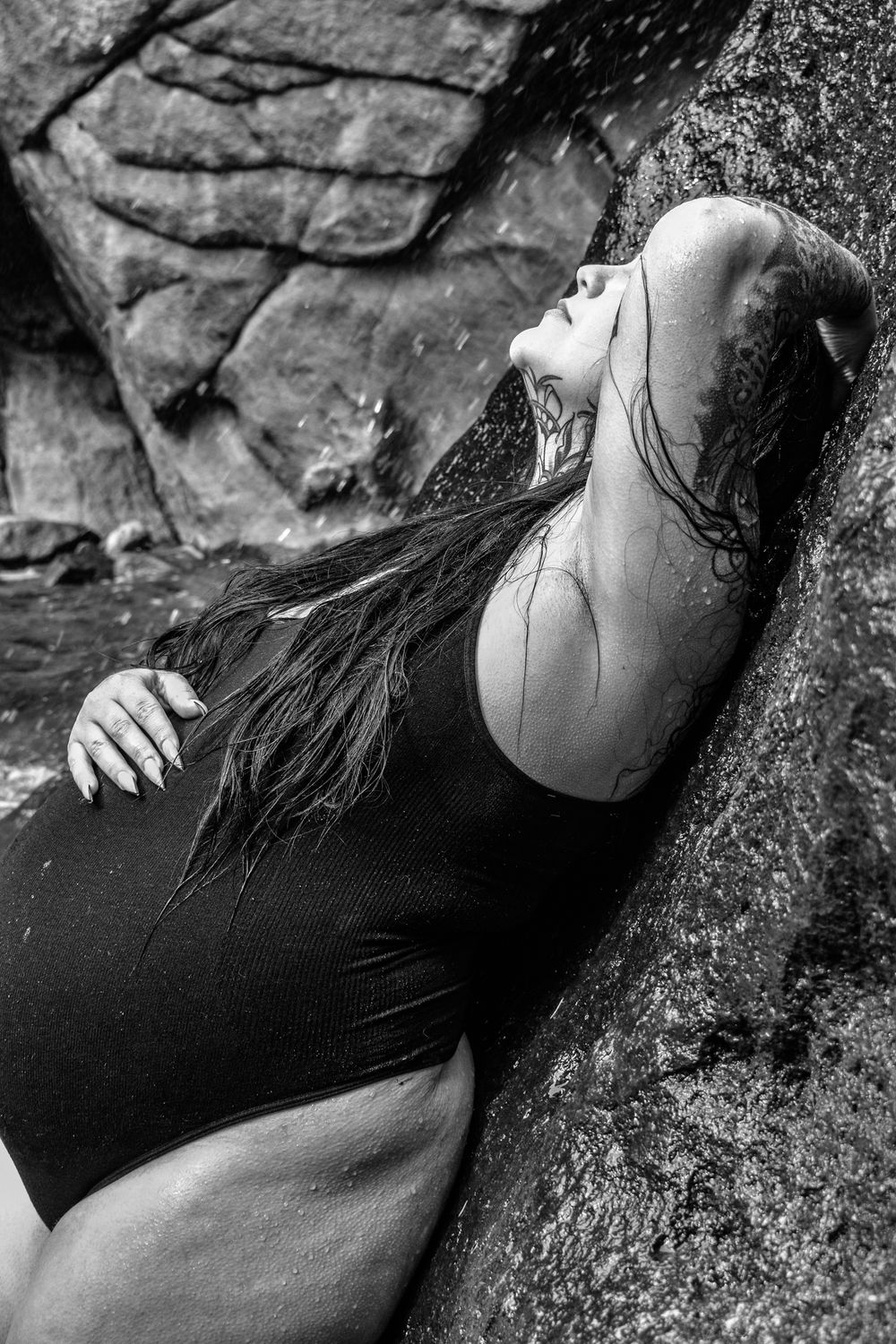 Woman in black swimsuit leans against wet rocks with long hair flowing in dramatic black and white photo.