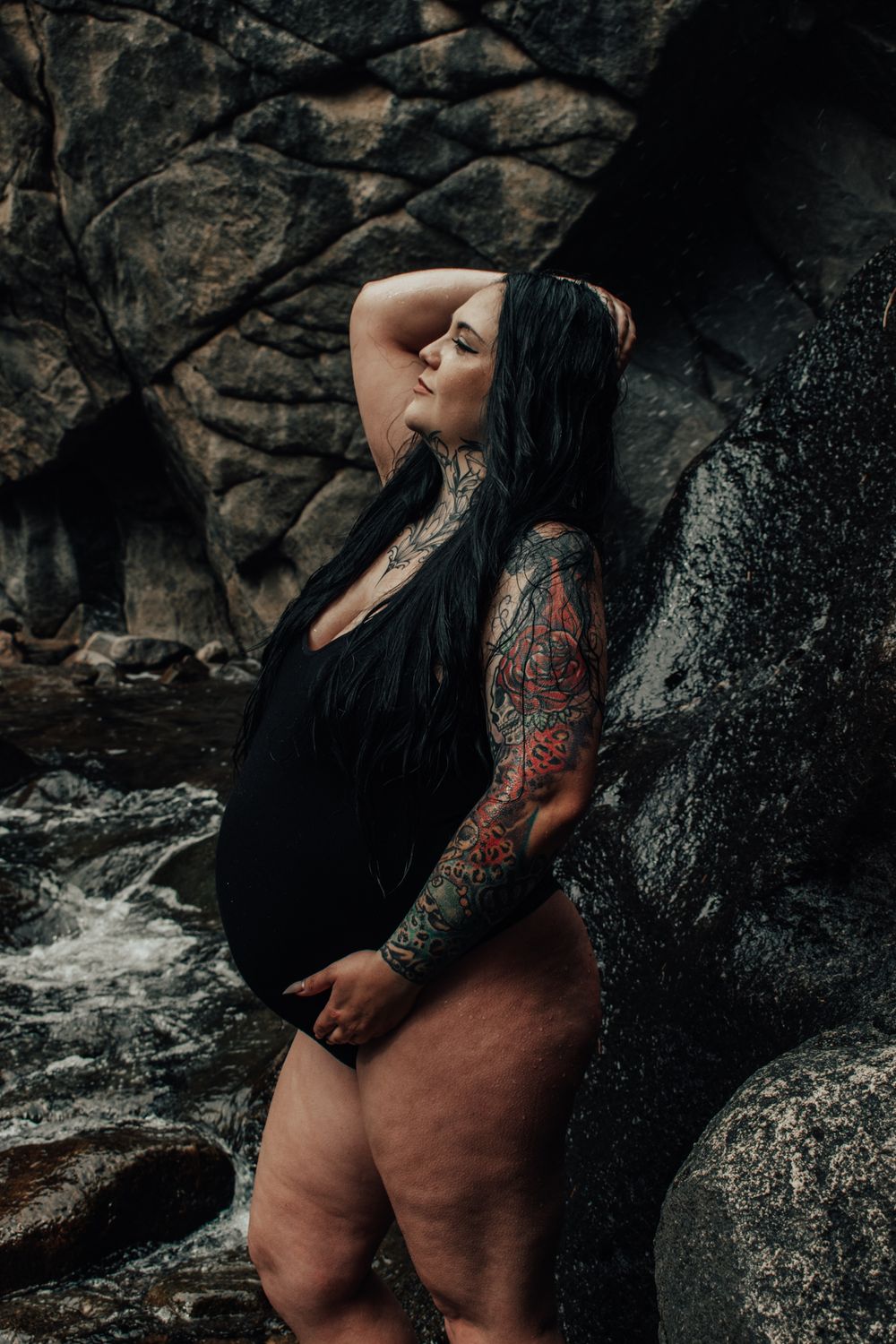 Pregnant woman with tattoos in black swimsuit poses against rocky backdrop near a stream.