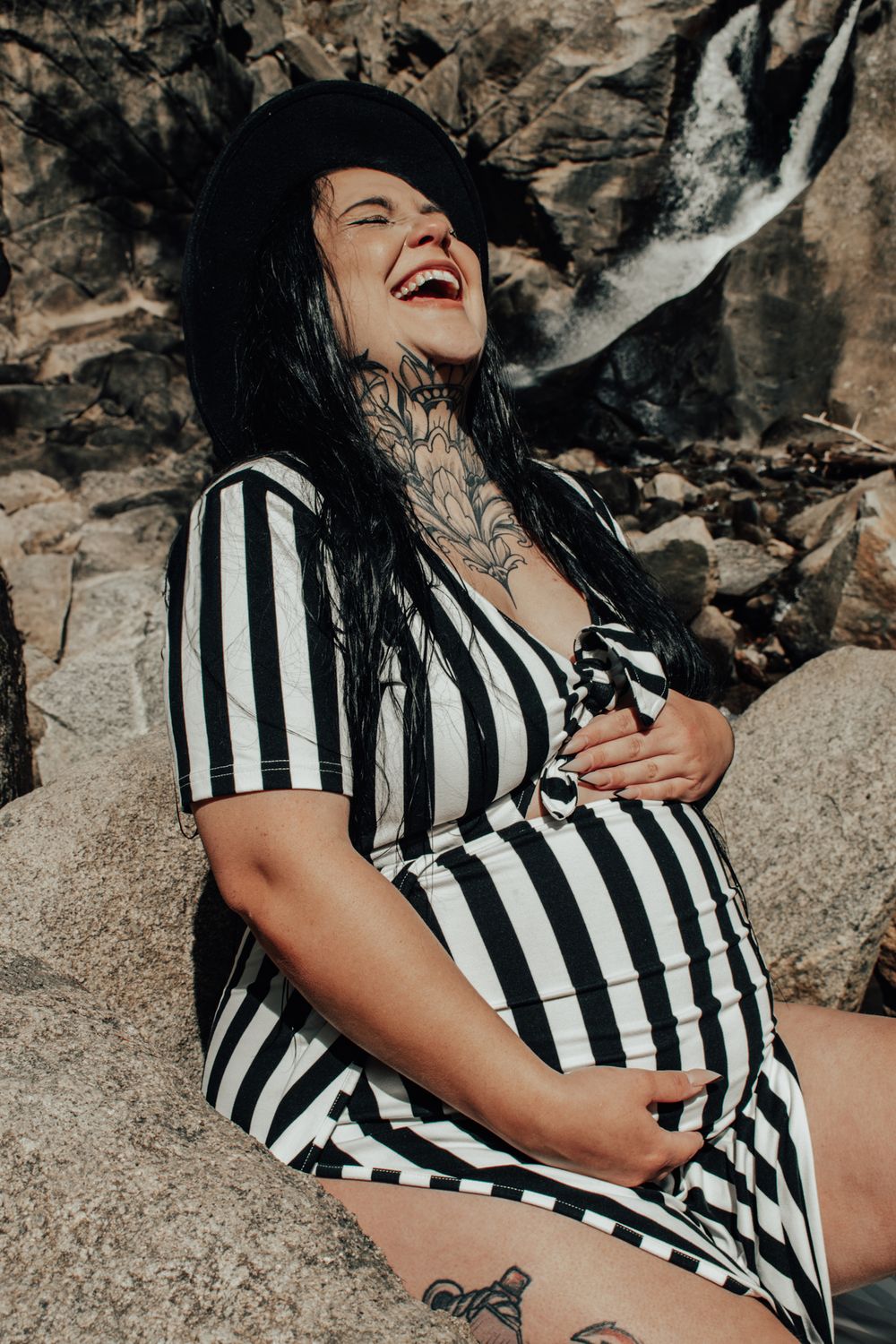 Laughing pregnant woman with tattoos in black and white striped dress sitting by rocky waterfall.