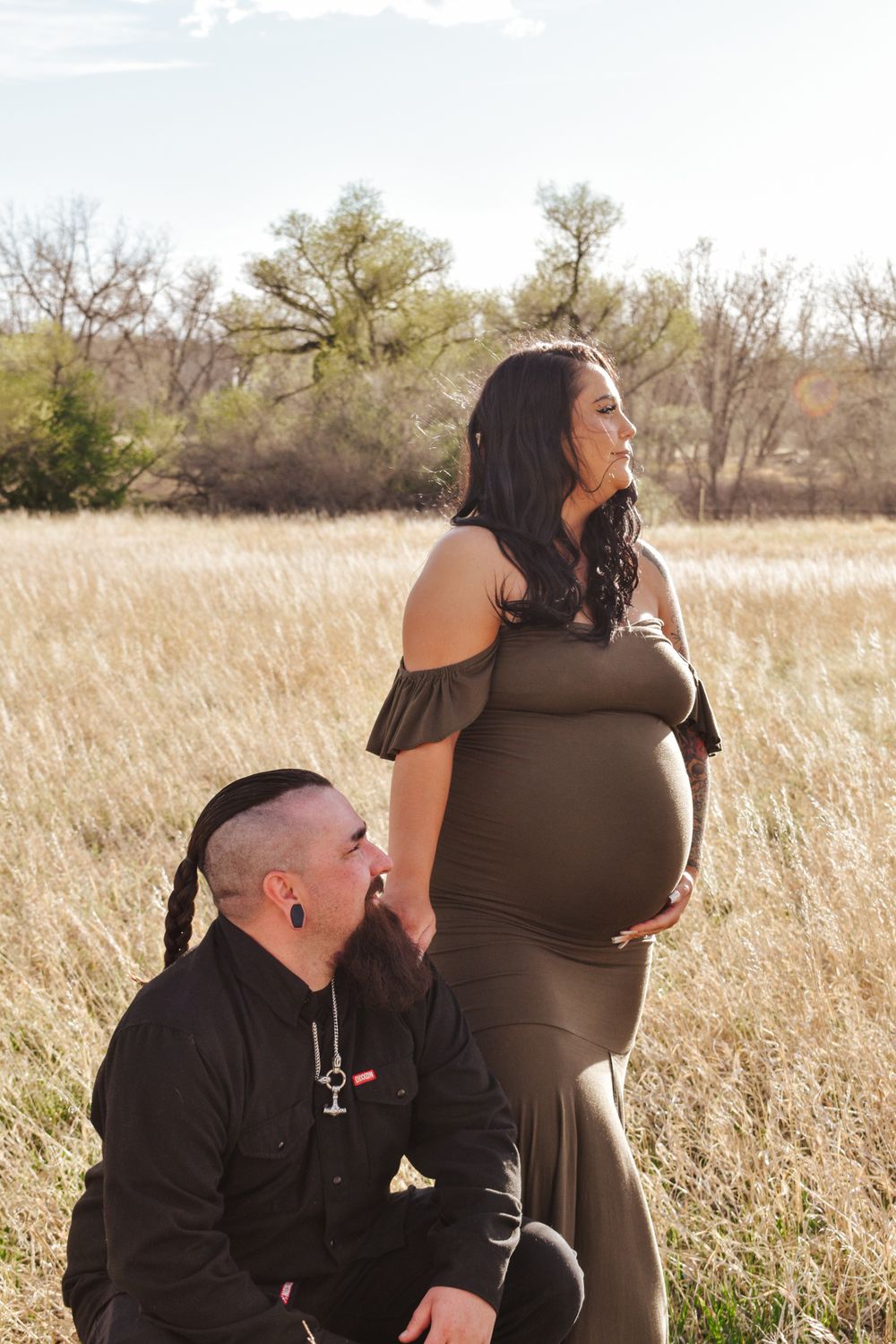 Pregnant woman in olive dress holds baby bump outdoors while partner kneels beside her in golden grass field.