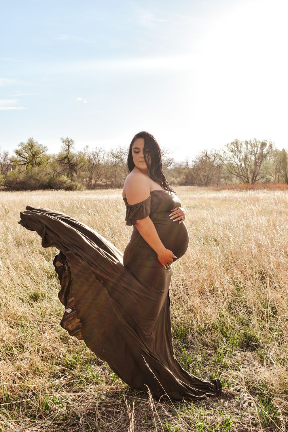 Pregnant woman in flowing olive off-shoulder gown poses in sunlit golden field with dress billowing in wind.