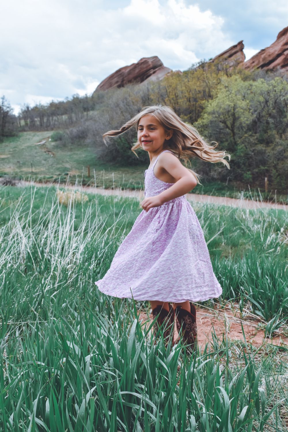 Young girl in purple dress twirling in green field with red rock formations in background.