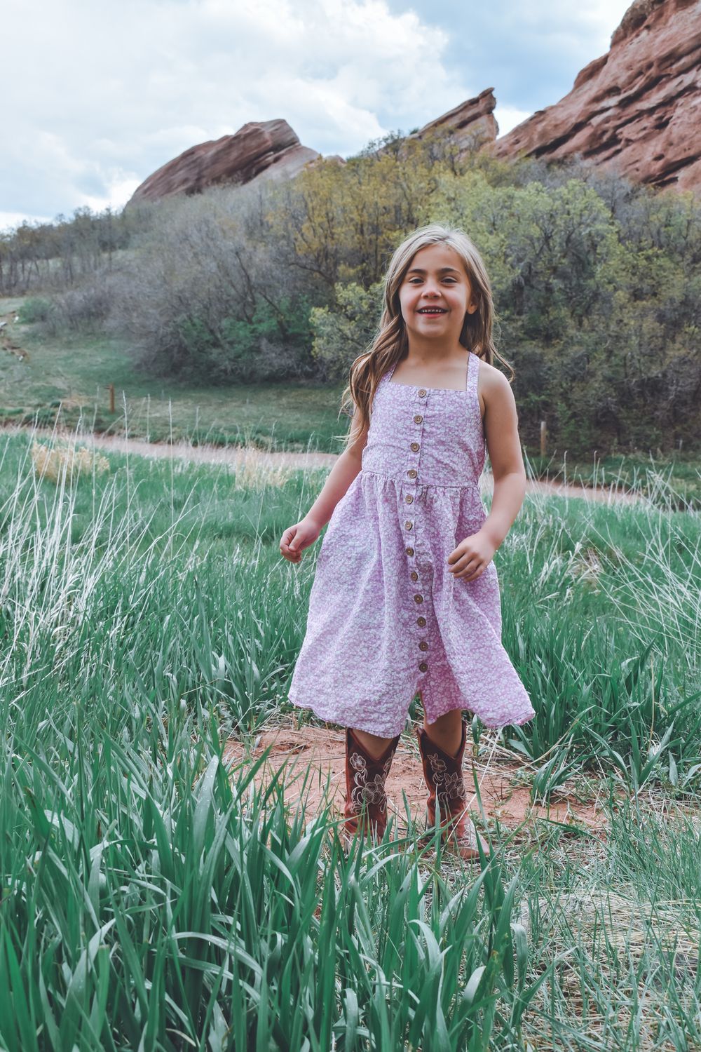 Young girl in pink dress and cowboy boots smiling in green field with red rock formations behind her.