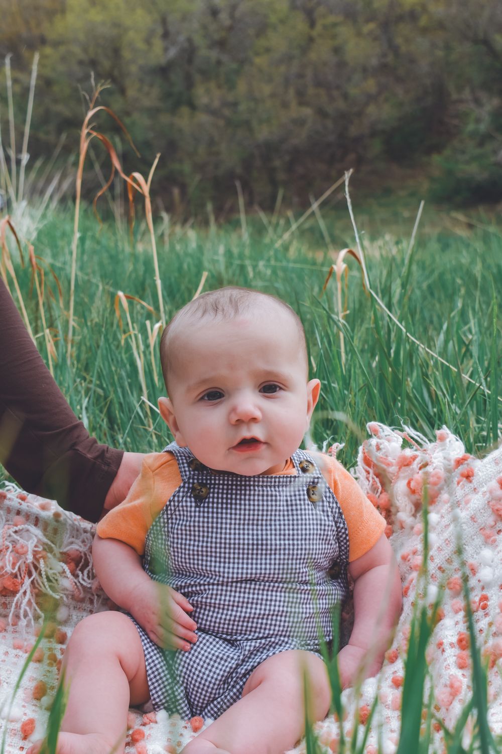 Adorable baby in checkered overalls sits on floral blanket surrounded by tall green grass outdoors.