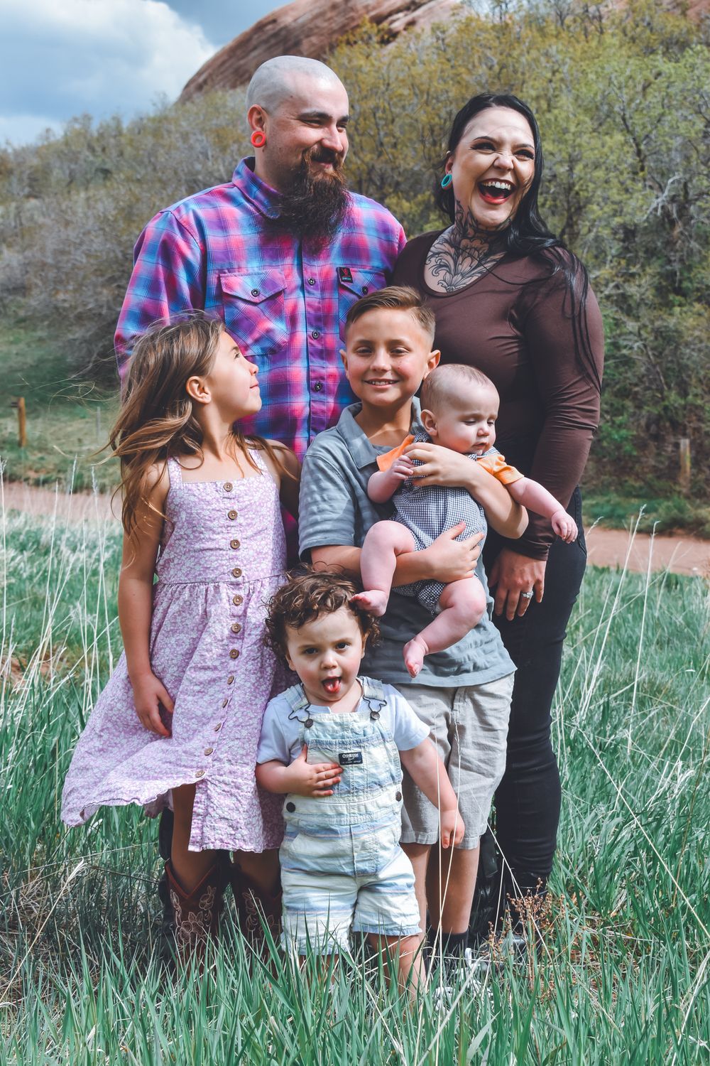 Happy family of six with parents and four children pose laughing in green meadow with red rocks backdrop.