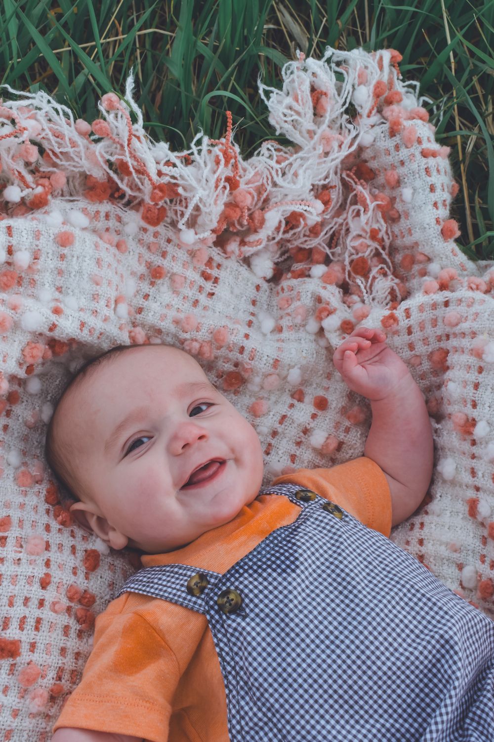 Smiling baby in gingham overalls lying on a textured blanket in the grass.