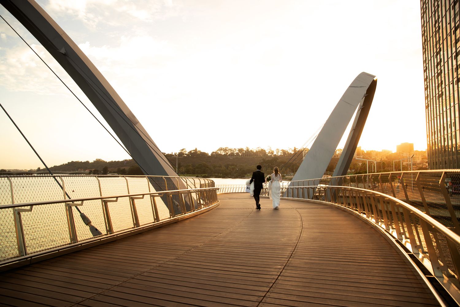 Person walking on a curved wooden pedestrian bridge at sunset with water and city views in the background.