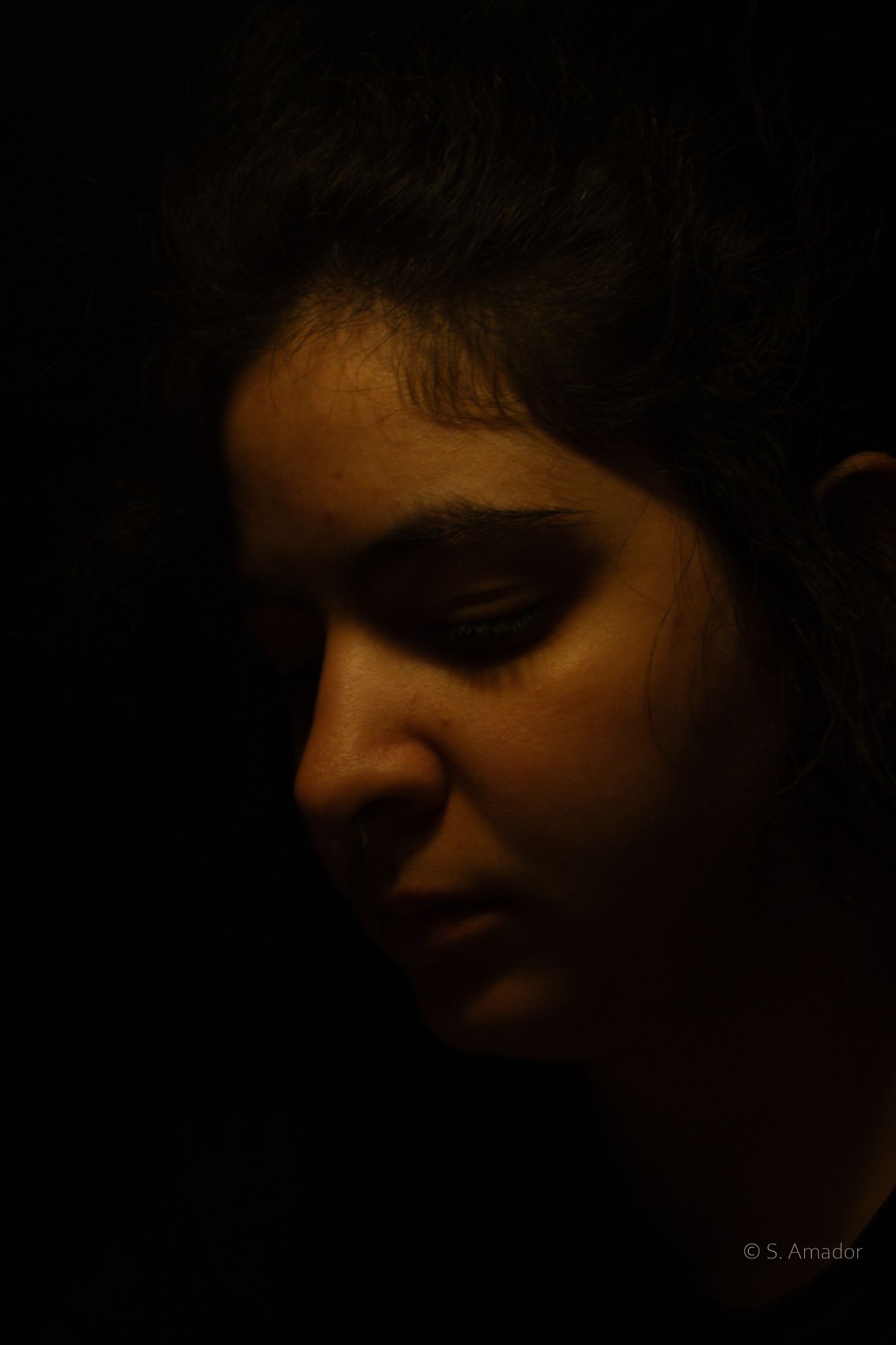 Dramatic low-key portrait of a young woman with eyes closed, illuminated by warm golden light against dark background.