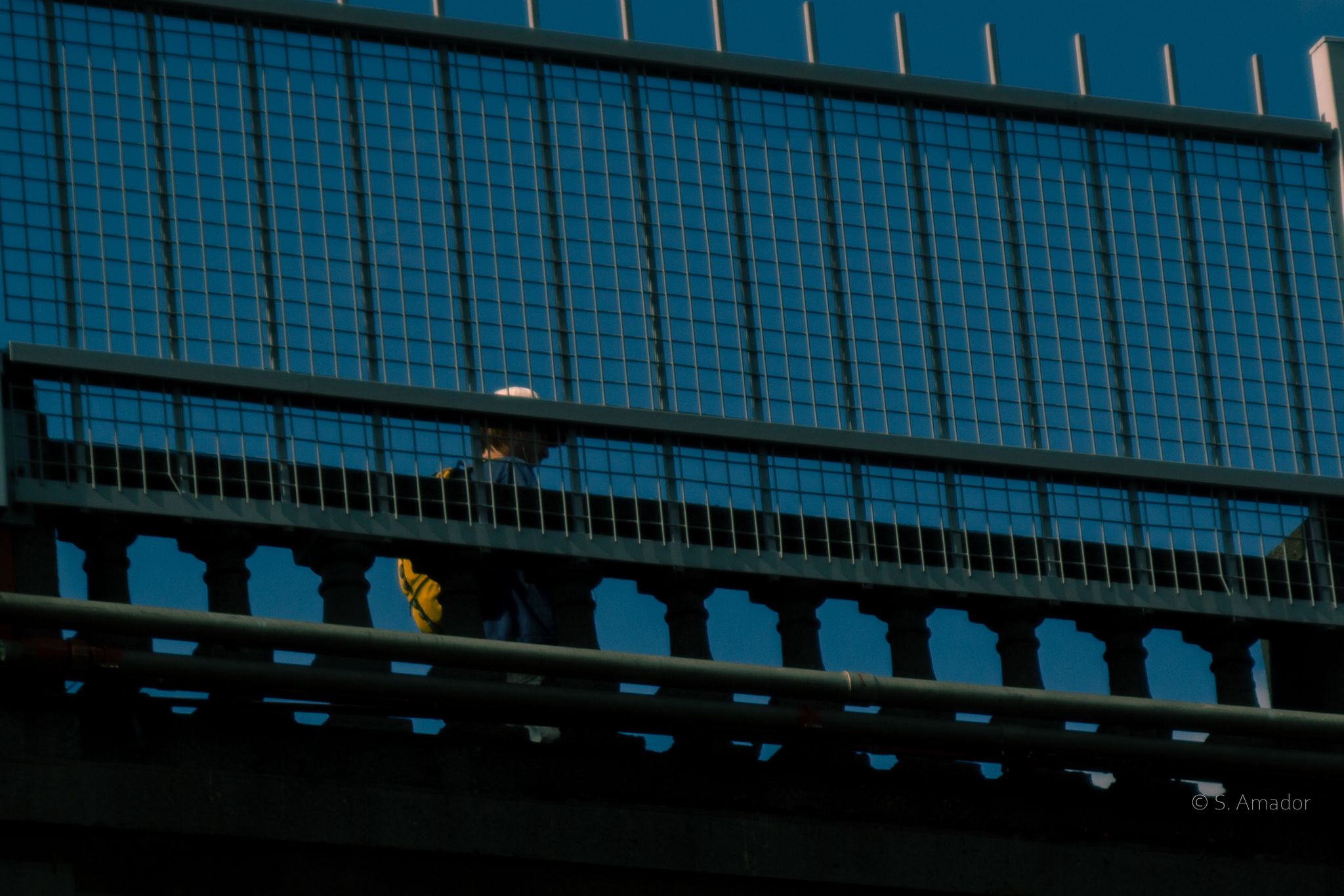 Person walking on a bridge overpass against a deep blue sky background in an urban setting.