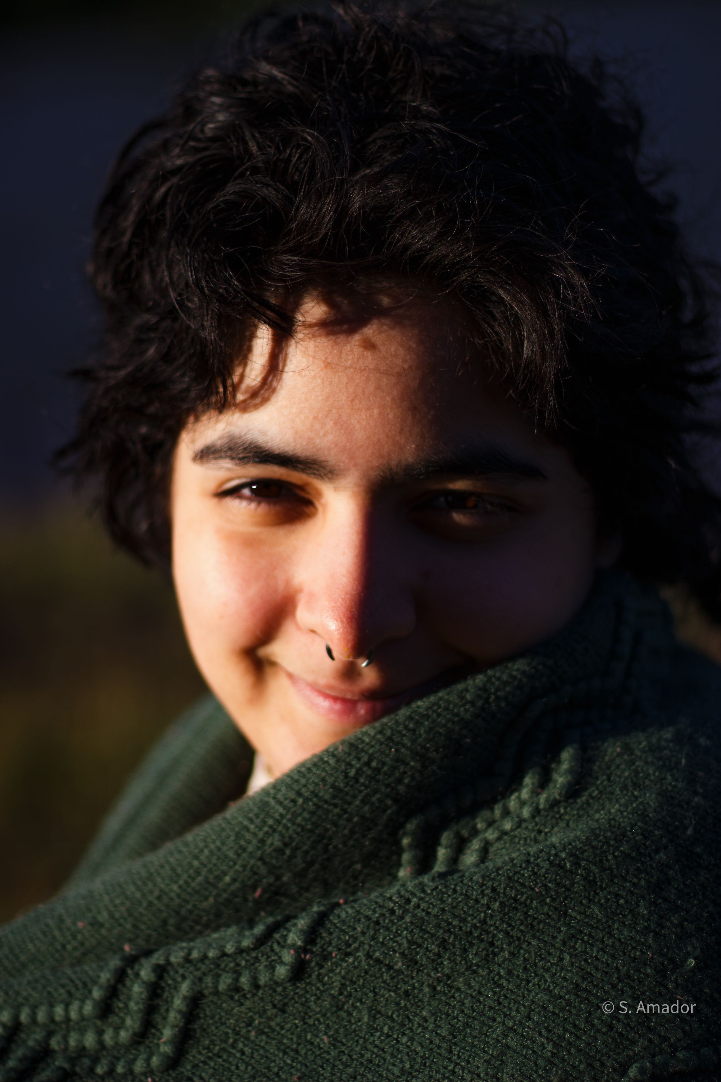 Young person with curly dark hair and nose ring smiling warmly, wrapped in a green knit sweater in natural light.