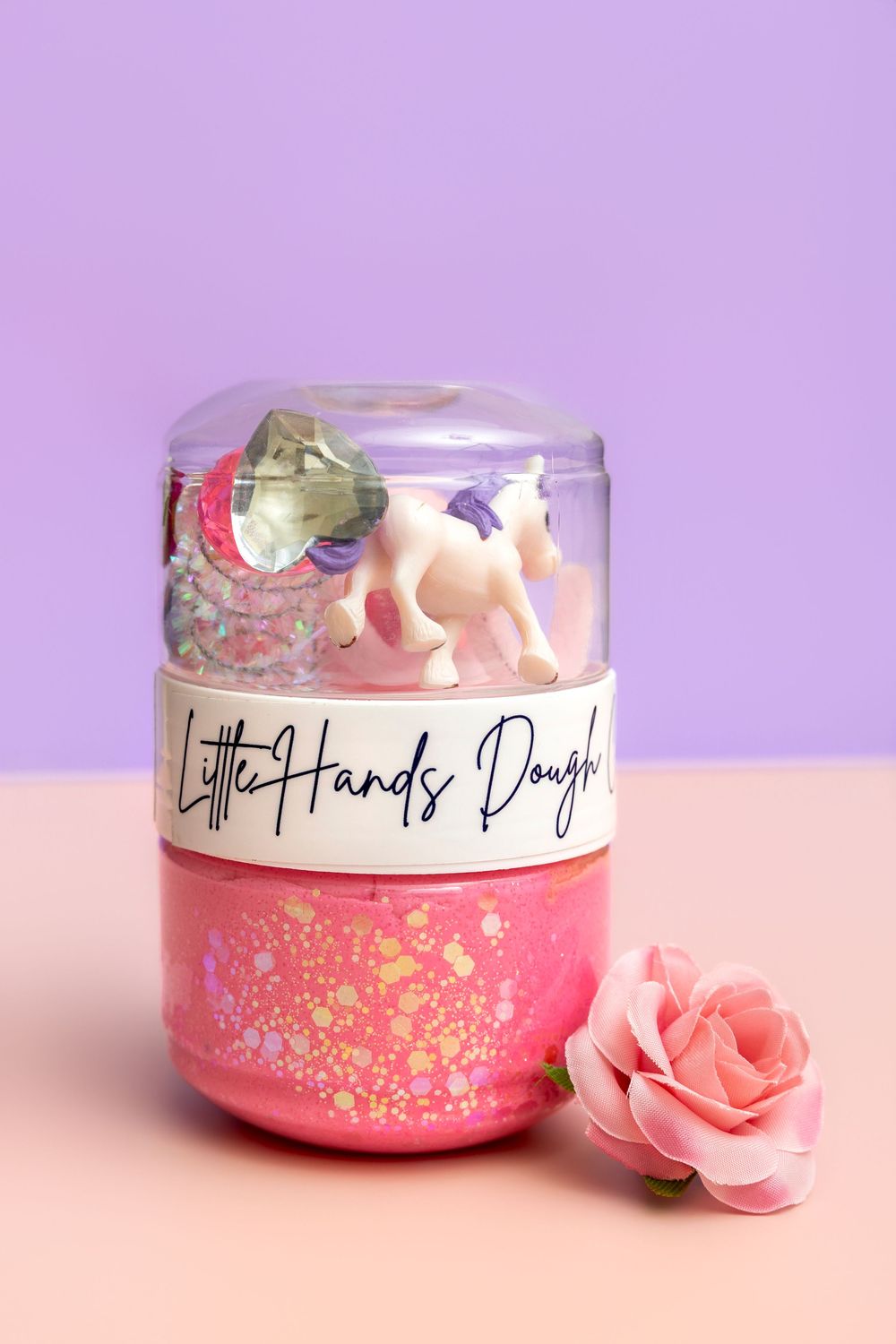 Pink glittery Little Hands Dough jar with unicorn toy and colorful sequins, displayed with a pink rose on pastel background.