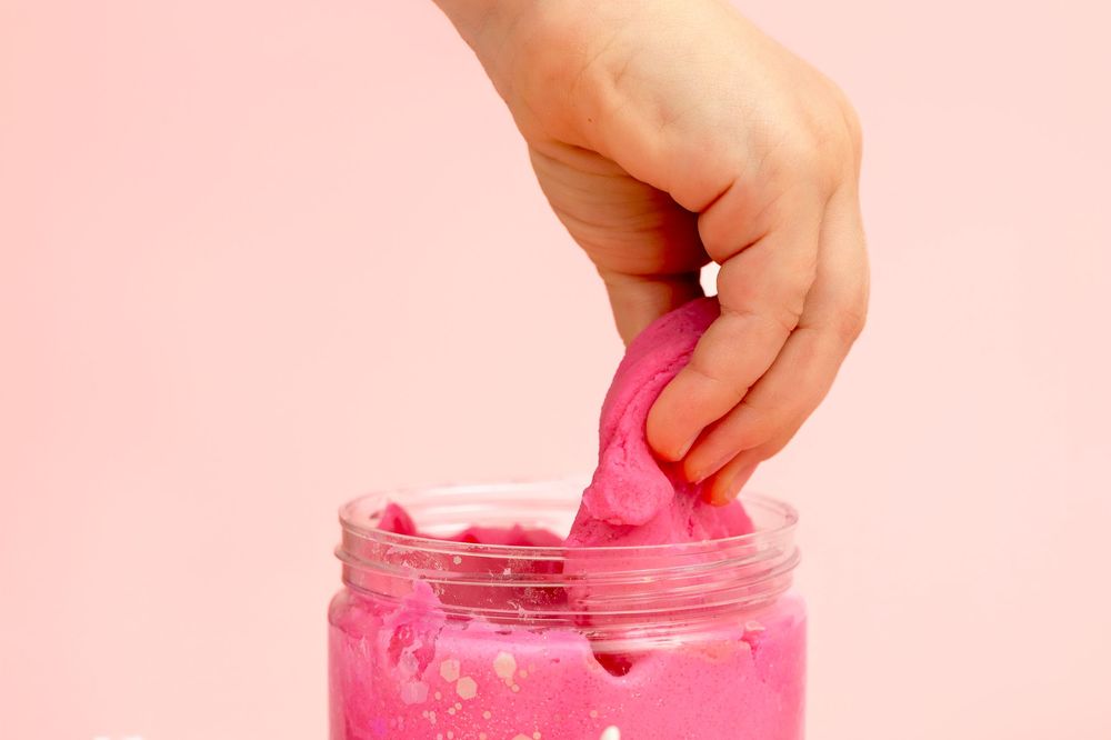 Hand pulling pink slime from a glass jar against a pink background.