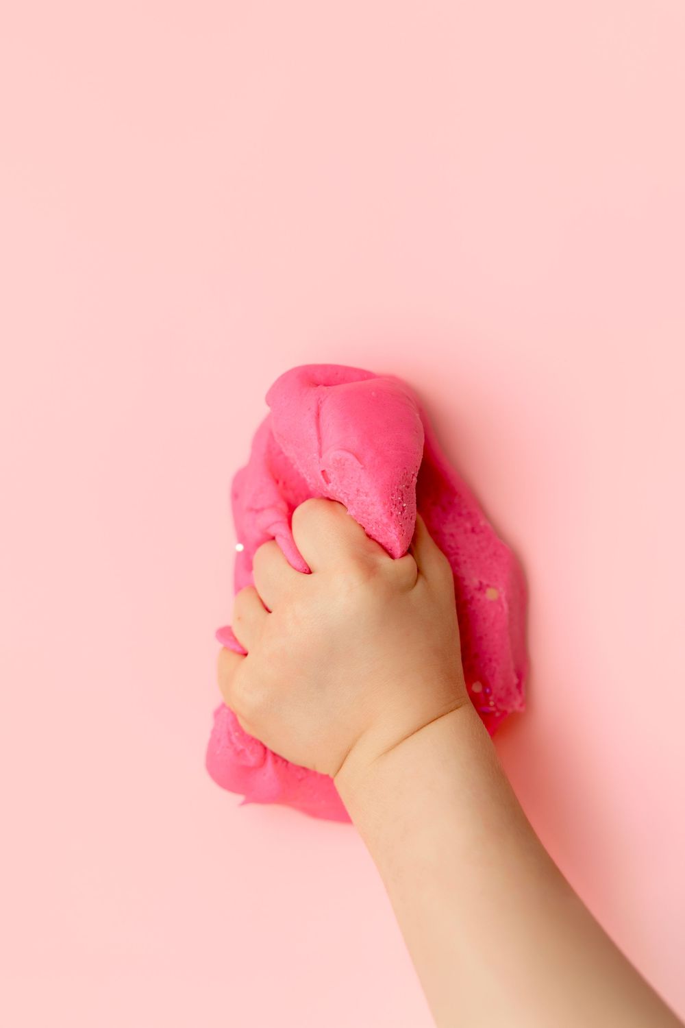 A child's hand squeezing bright pink slime or play dough against a soft pink background.
