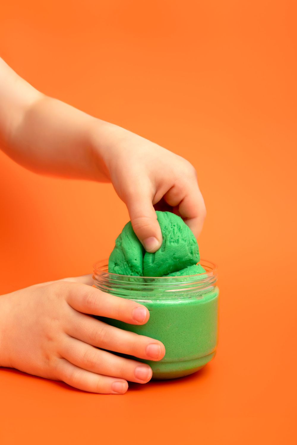 Child's hands playing with green play dough from an open jar on a bright orange background.