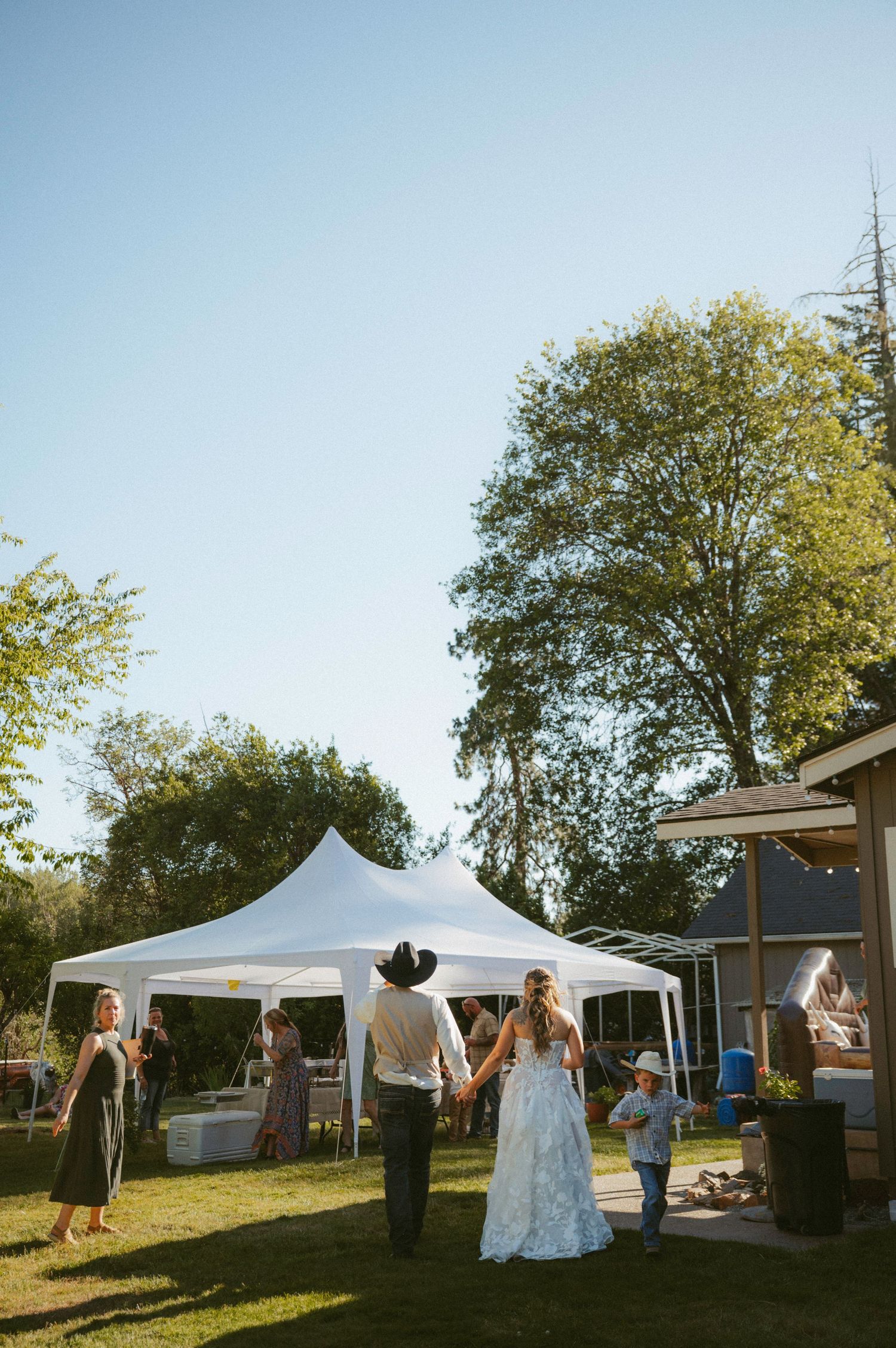Bride and groom hold hands outdoors at a backyard wedding reception with white tents on a sunny day.