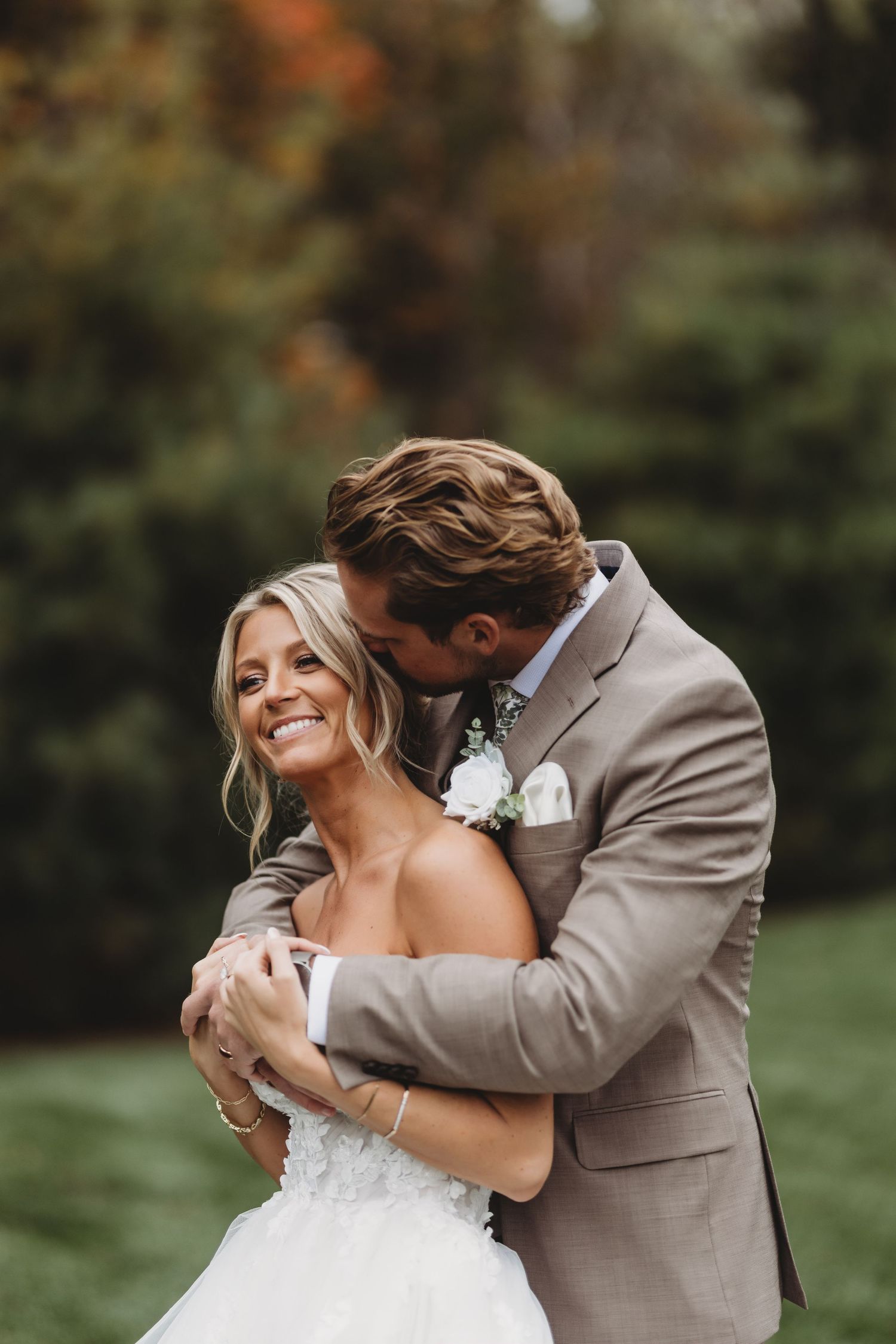 A smiling bride in a white gown embraced by a groom in a tan suit, sharing a tender moment outdoors.