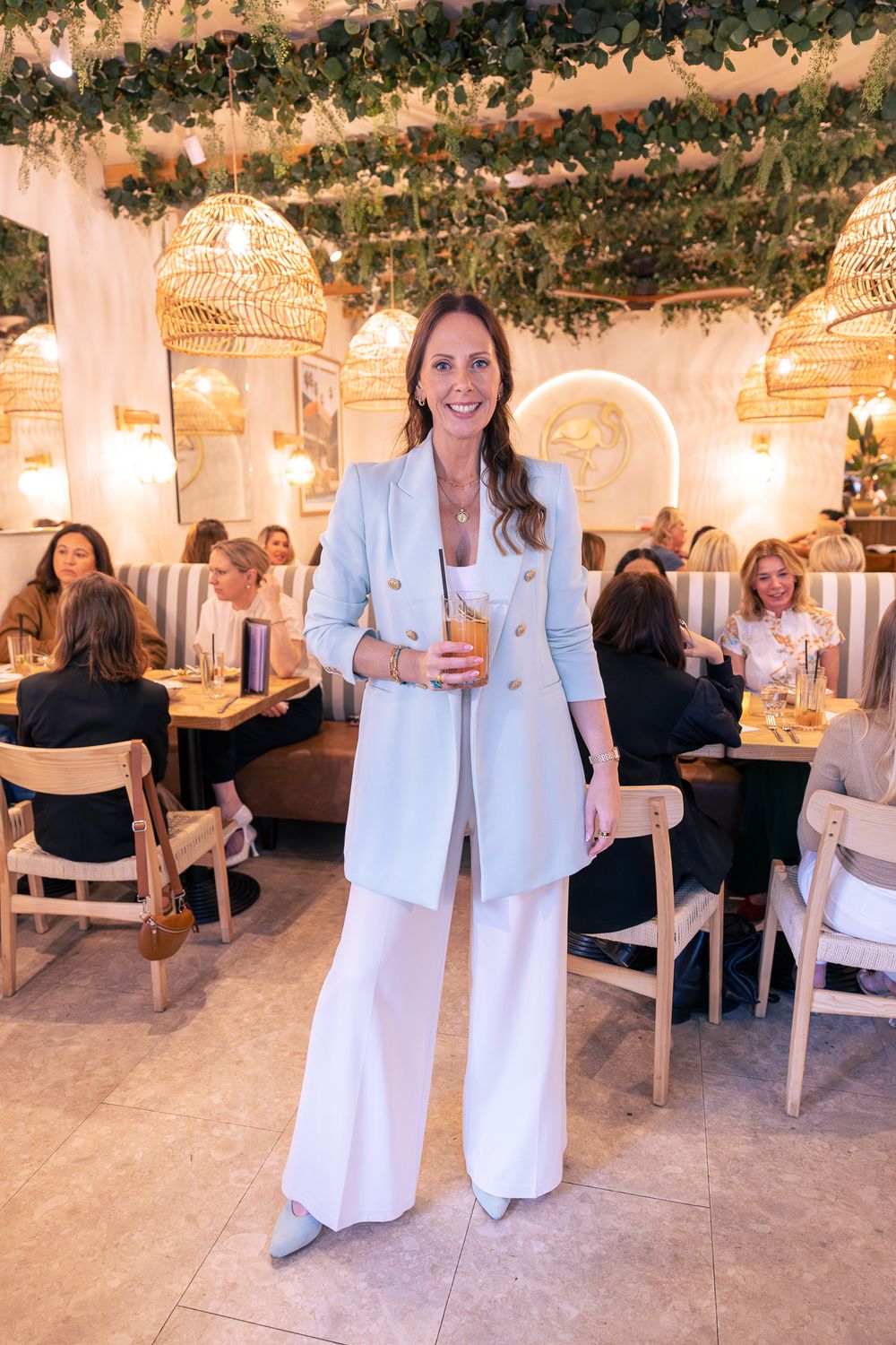 Woman in light blue suit holding cocktail stands in elegant restaurant with wicker pendant lights and greenery.