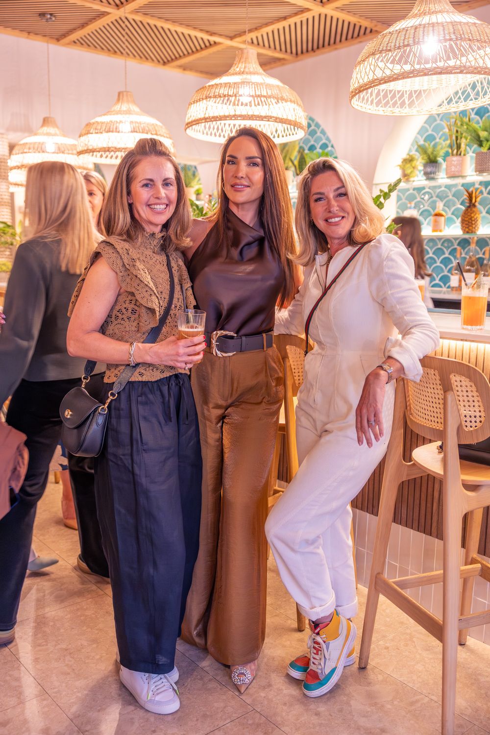Three stylish women posing together at a chic restaurant with rattan pendant lights and blue tile accents.