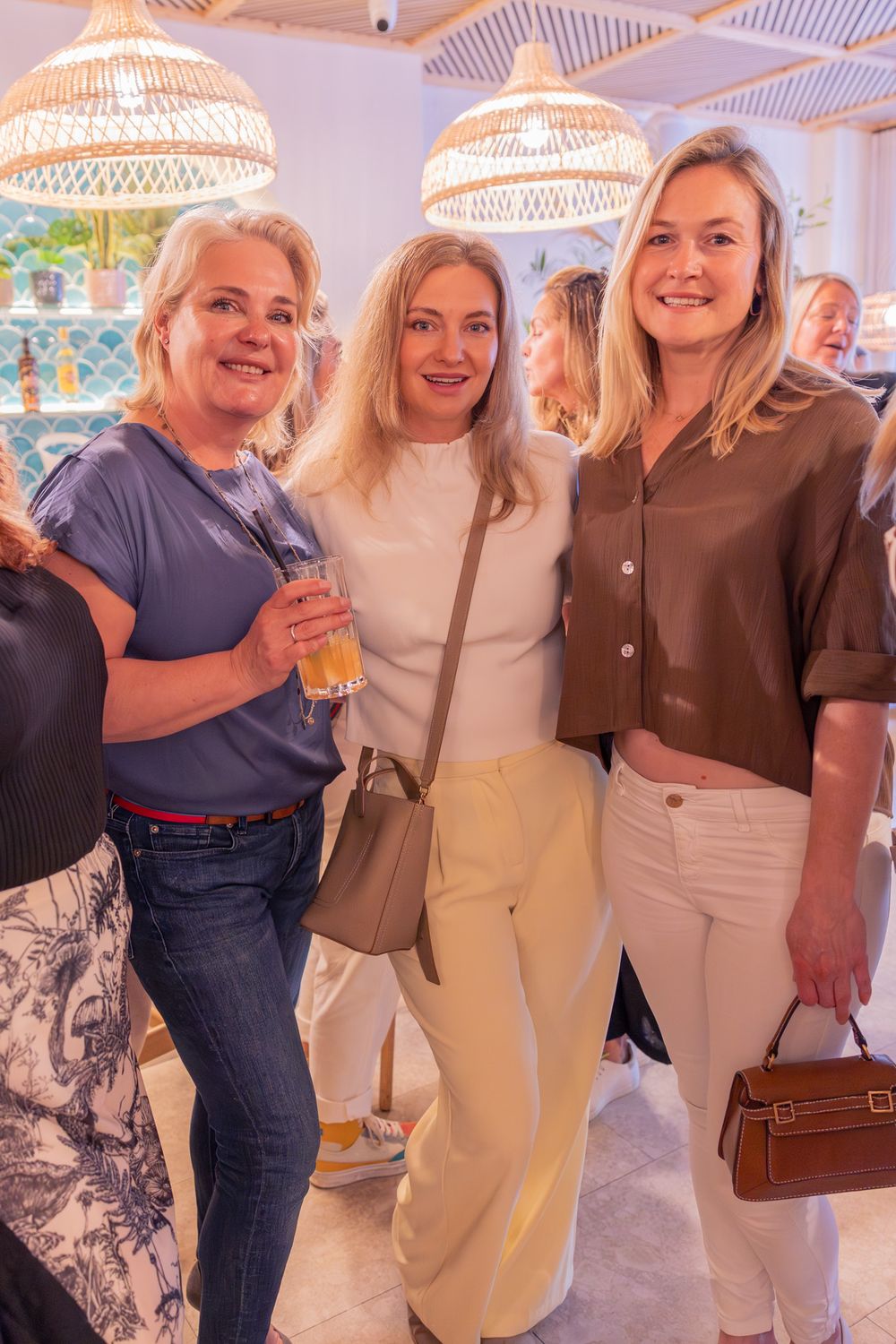 Three smiling women posing together at a social event, dressed in casual chic outfits, holding drinks and handbags.