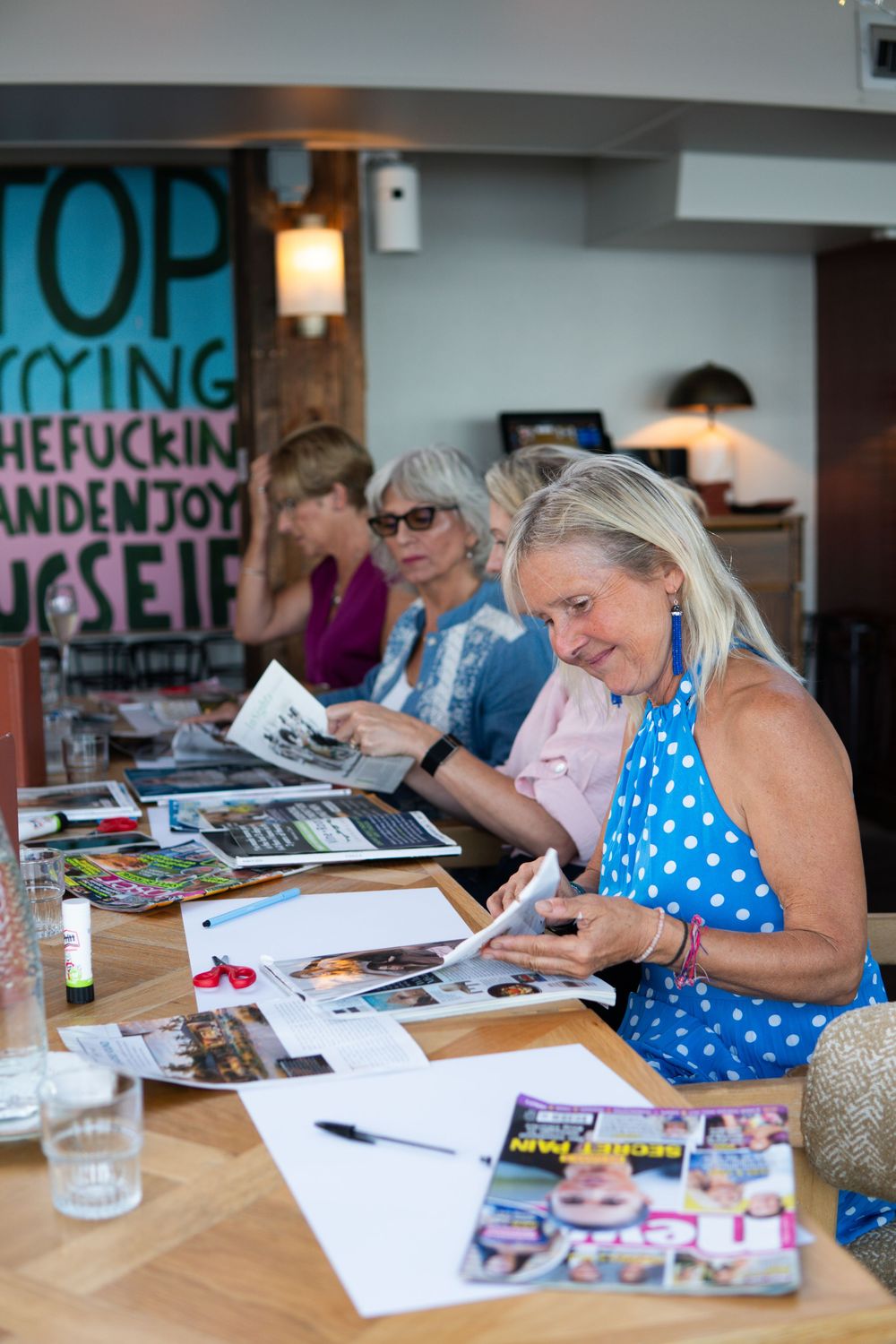 Women sitting at a table engaged in a creative collage workshop, cutting and arranging magazine images indoors.