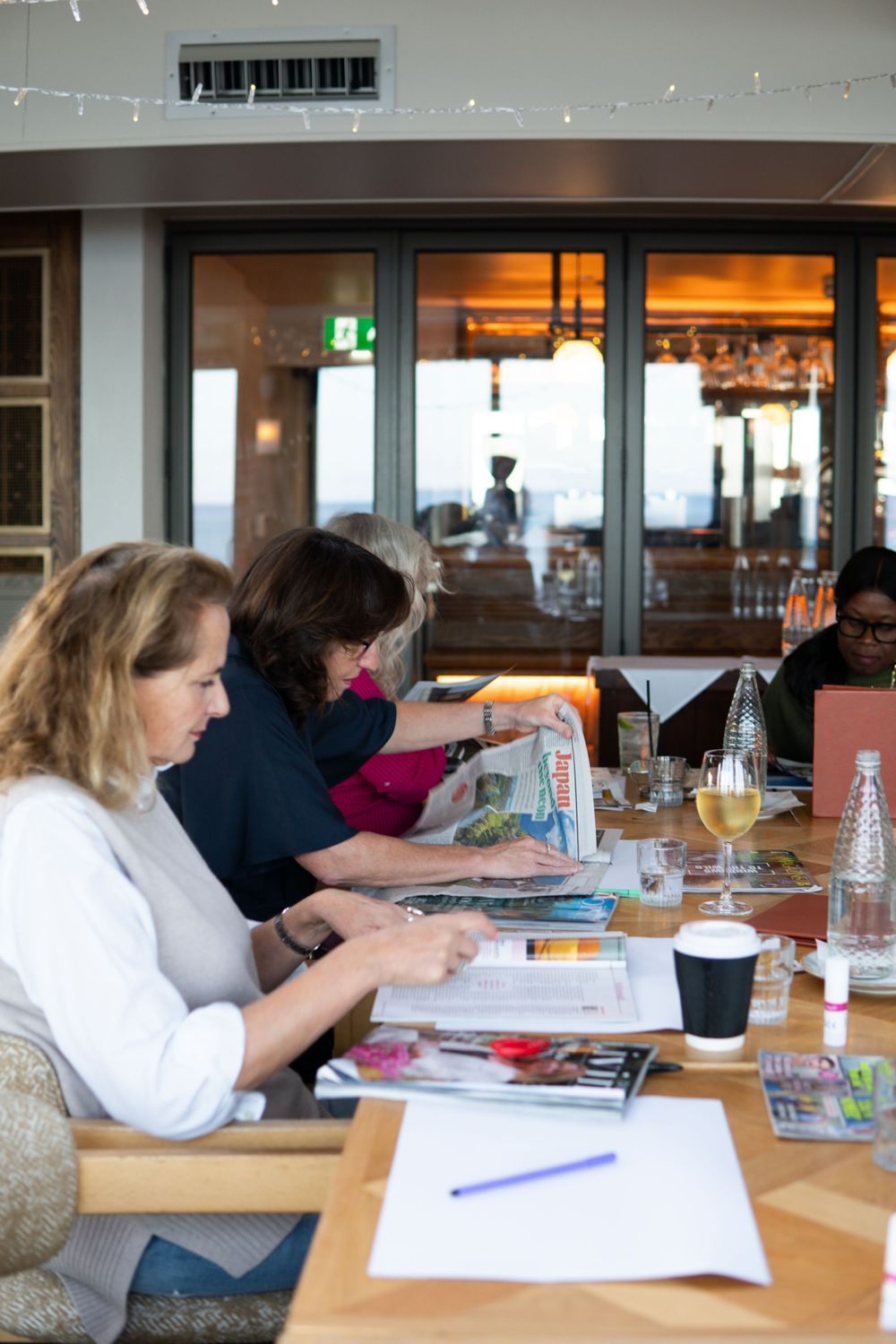 Women sitting at a long table in a restaurant, reading magazines and newspapers with drinks nearby.