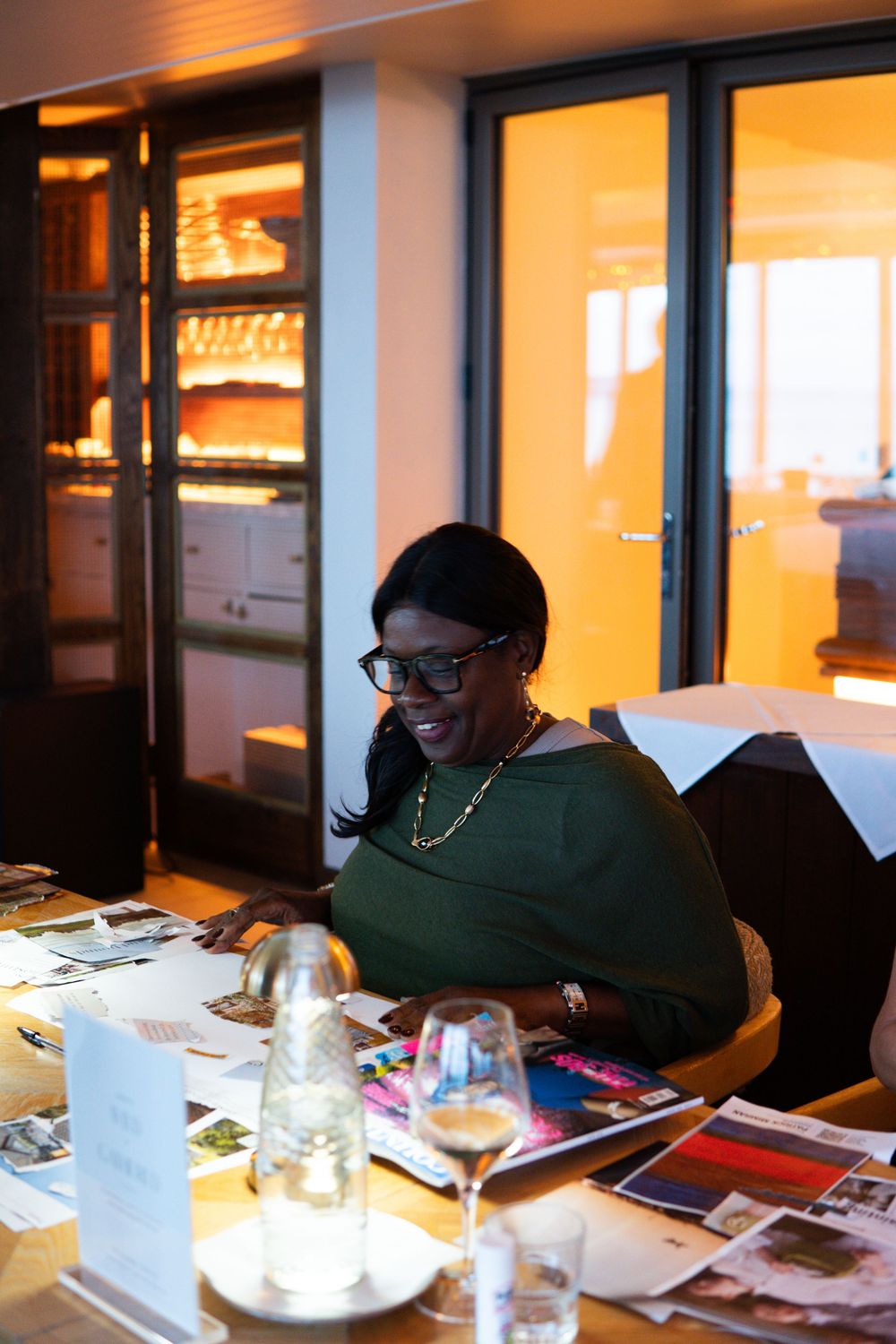 Woman in green top and glasses reviews magazines at a restaurant table with wine glasses and warm lighting.