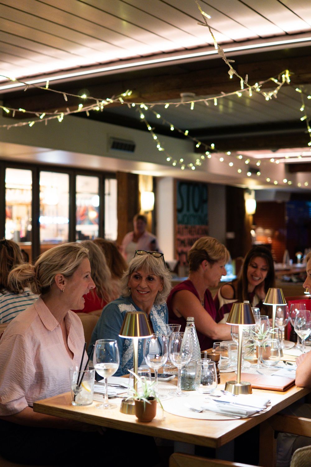 Women dining at a restaurant table with fairy lights, wine glasses, and warm ambiance.
