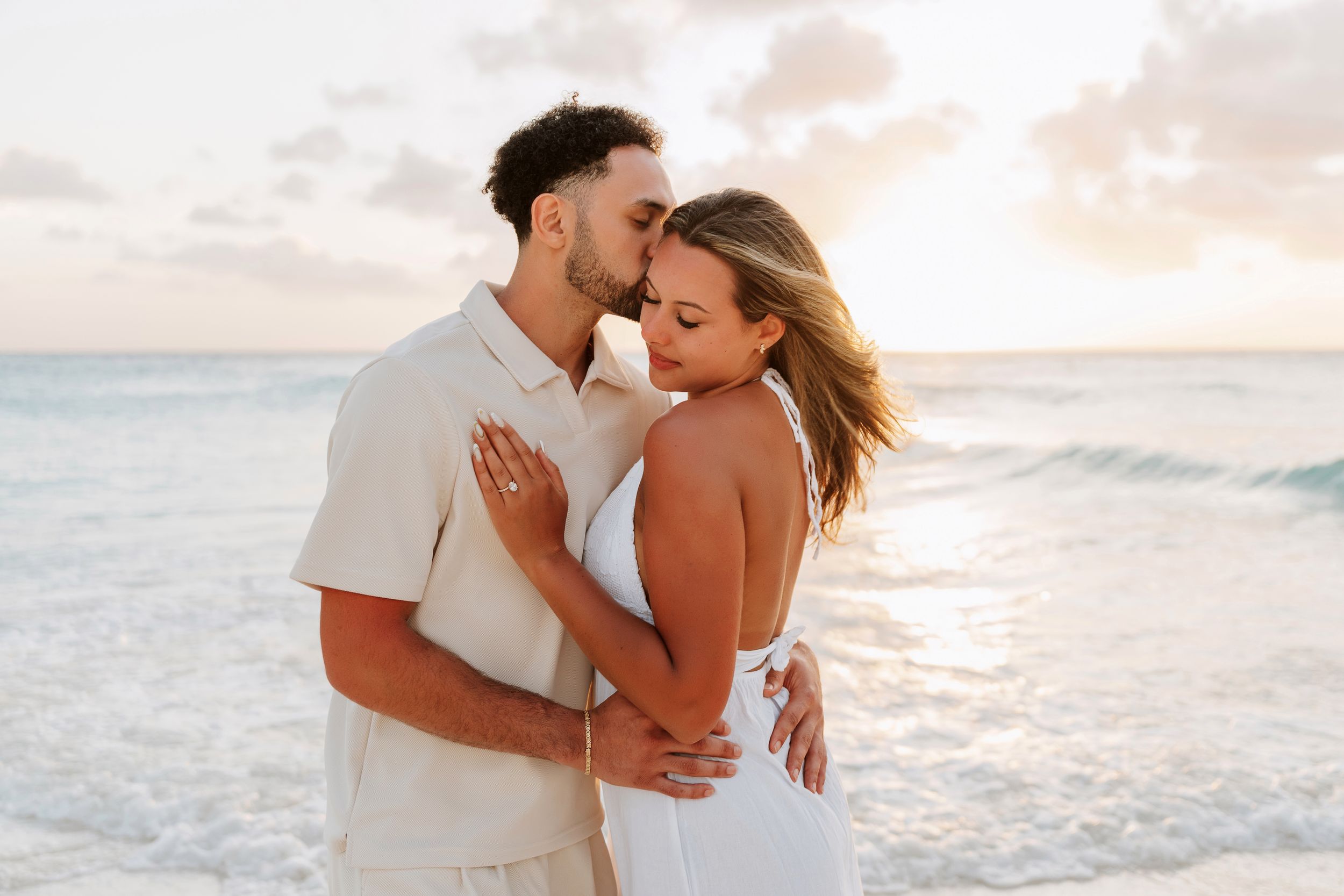 Couple holding hands at sunset beach proposal setup with floral arch, candles, and Marry Me sign on sandy shore.