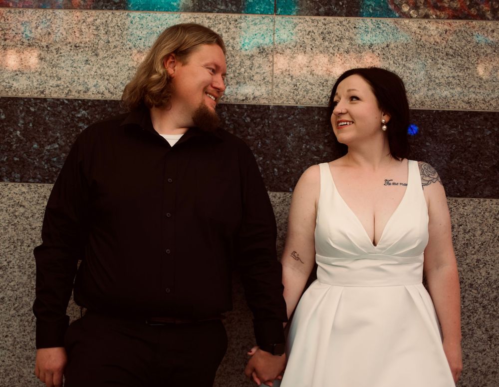 A couple in wedding attire holds hands and smiles at each other against a decorative wall backdrop.