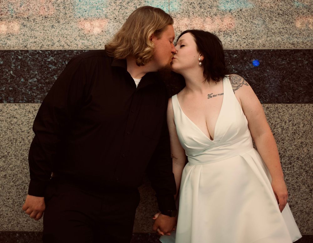 Couple sharing a romantic kiss at their wedding, bride in white dress with tattoo, groom in black suit.