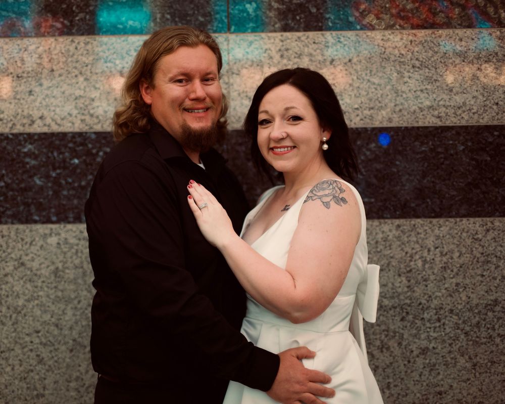 A smiling couple in wedding attire poses together, the bride in white with a shoulder tattoo and groom in black.
