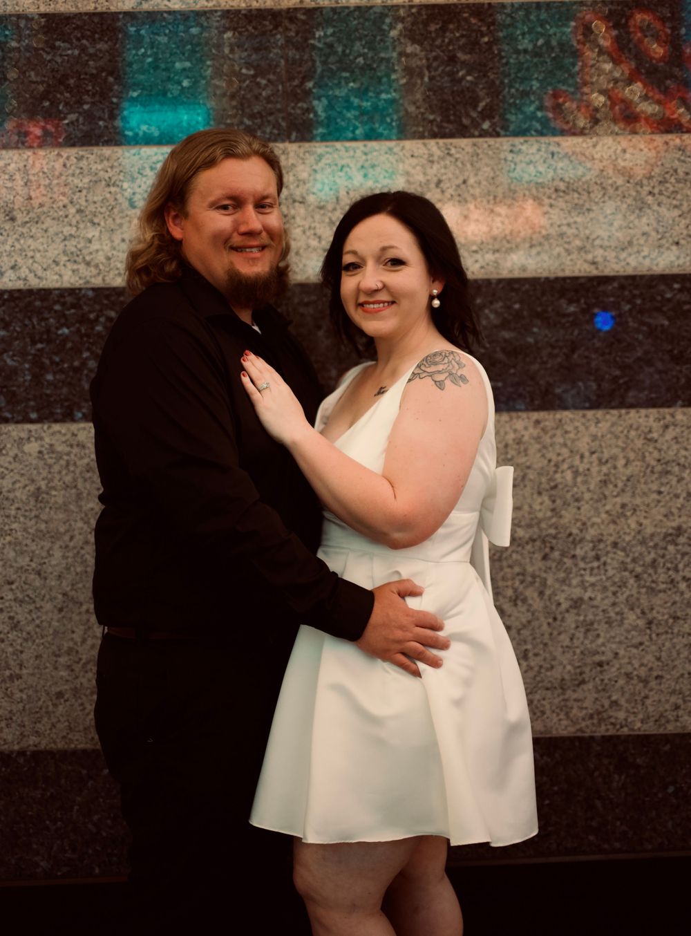 Couple posing together, man in black suit and woman in white mini dress, smiling at wedding celebration.