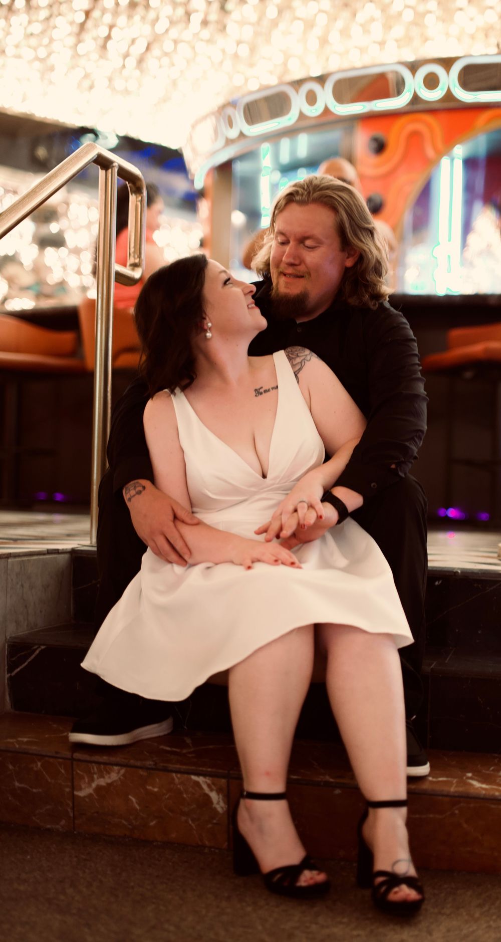 Smiling bride in white dress sitting on steps with groom behind her in glittering casino venue.