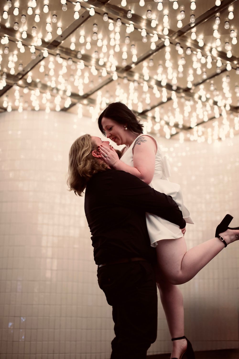 Groom lifting smiling bride under a ceiling of warm Edison bulb string lights.