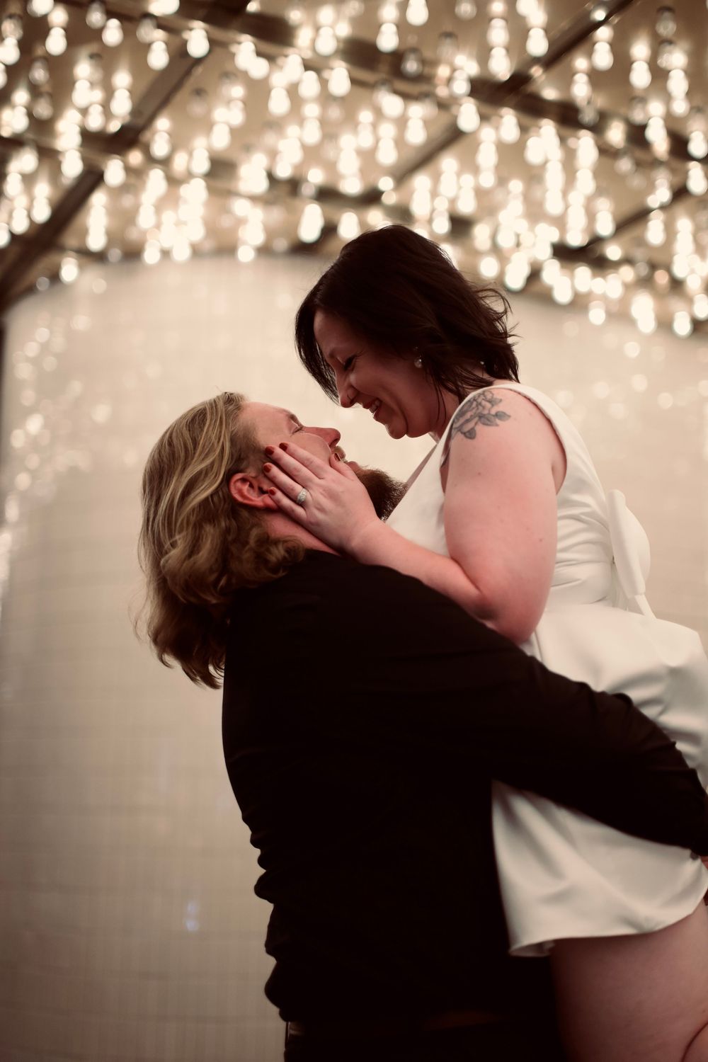 Romantic couple embracing and gazing at each other under warm bokeh string lights indoors.