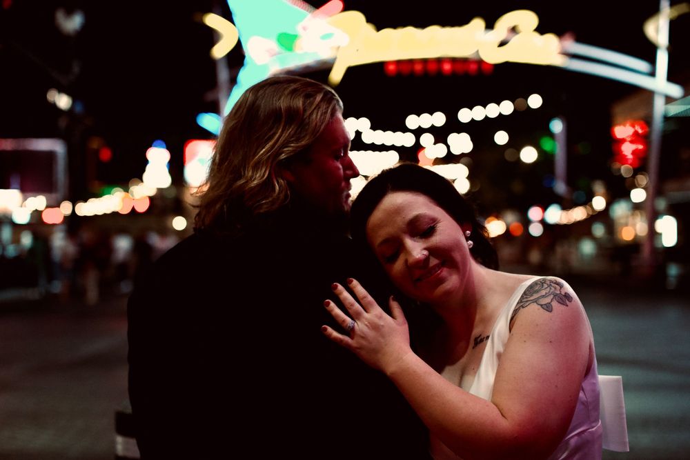 Couple embracing romantically at night on a bright neon-lit Las Vegas street, woman in white dress with tattoo smiling.