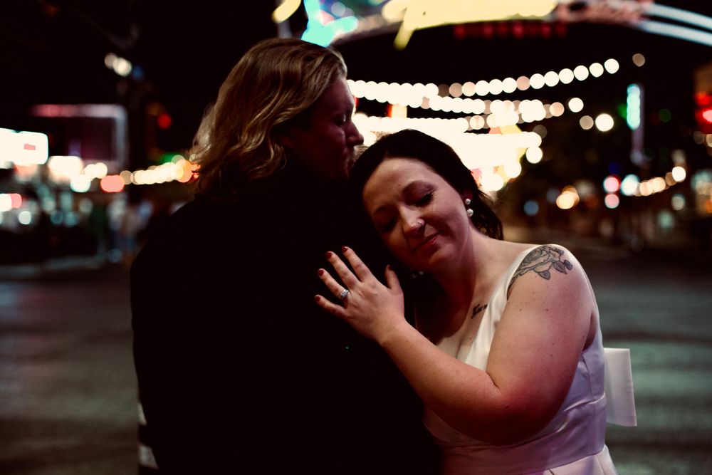 Couple embracing at night in a city street, bride in white dress with tattoo, groom in dark jacket, bokeh lights.